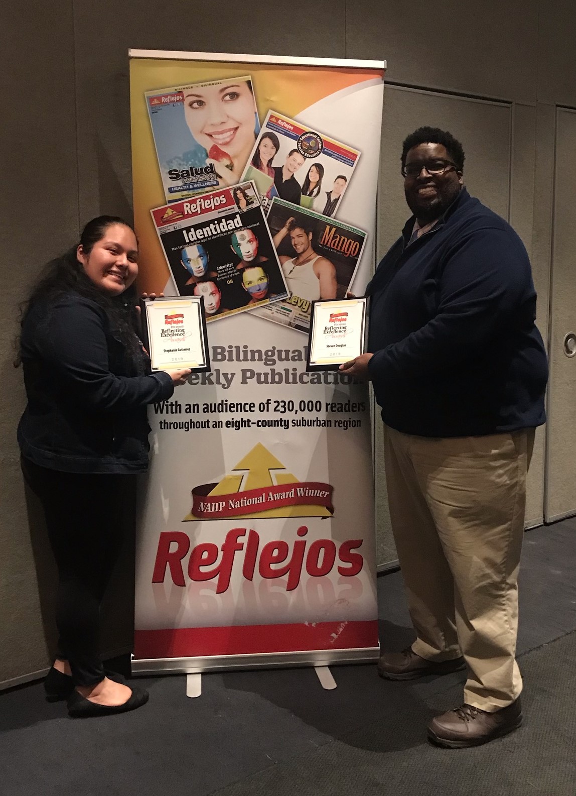 The two honorees standing in front of a banner for the magazine.